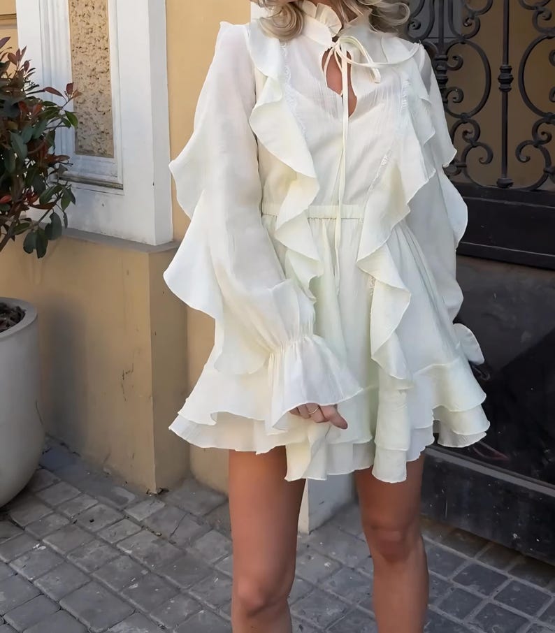 May include: A cream-colored mini dress featuring long sleeves with flared cuffs, a high ruffled collar with a tie closure, and vertical ruffles cascading down the front. The garment has a cinched waist and a layered, tiered skirt design.