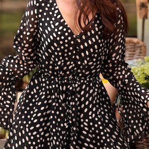 May include: Black romper with white polka dots. The V-neck romper features long, flared sleeves and a cinched waist. The fabric appears to be a lightweight, semi-sheer material. The overall style is a playful, yet elegant design.
