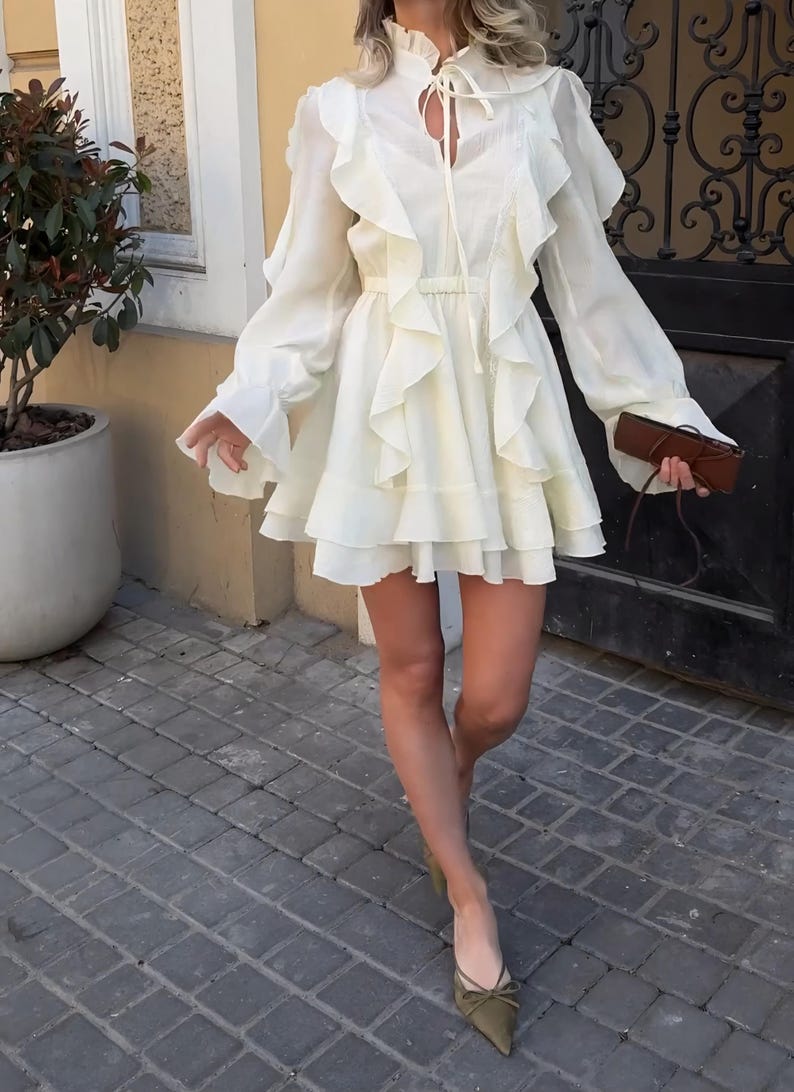 May include: A cream-colored mini dress featuring long sleeves with ruffled cuffs, a high ruffled collar with a tie-front closure, and vertical ruffles cascading down the bodice. The skirt is flared with a tiered hemline and a cinched waist.