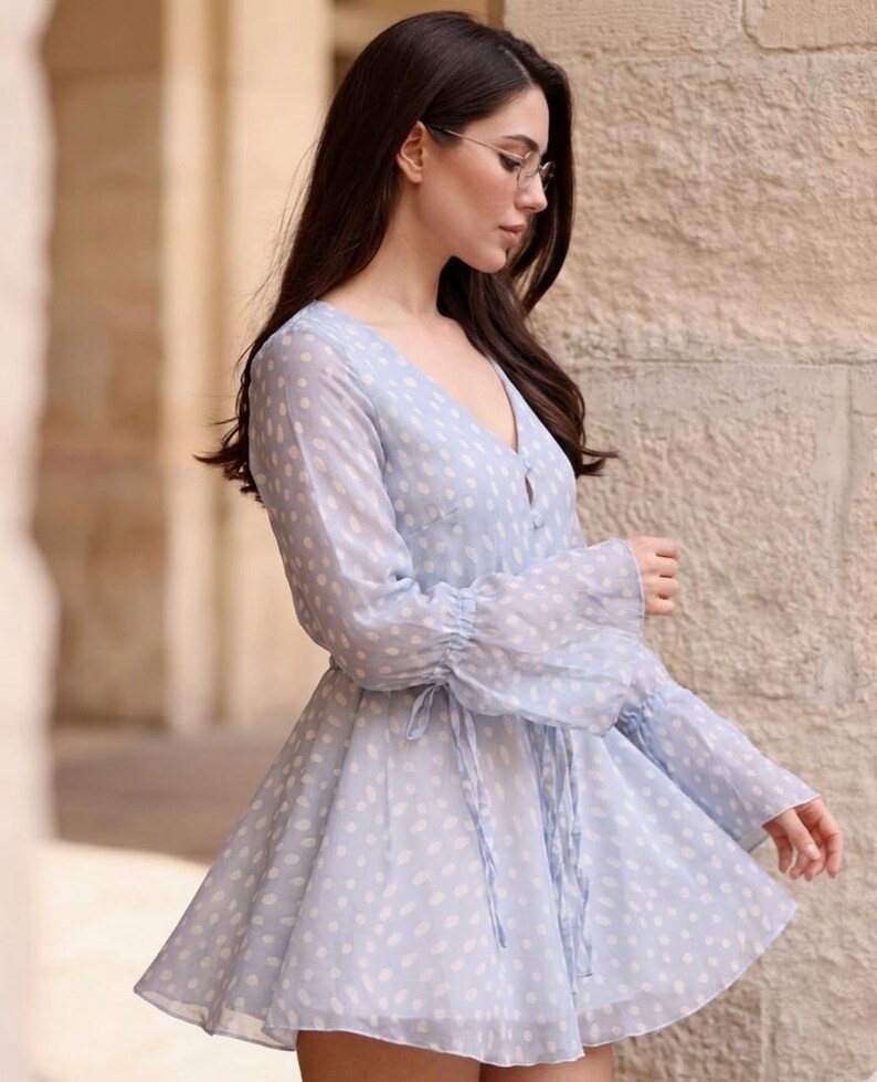 May include: A pale blue dress with white polka dots. The dress has long, sheer sleeves with frilled cuffs and a V-neck. A tie defines the waist, and the skirt flares. The dress is made of a lightweight material.