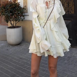 May include: A cream-colored long-sleeve dress featuring a high ruffled collar with a tie-front closure, vertical cascading ruffles along the bodice and sleeves, and a multi-layered flared skirt with a thin brown shoulder strap visible across the chest.