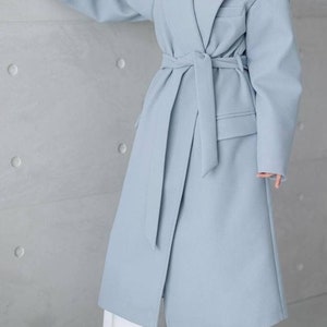 May include: A light blue coat with a wrap-around design and a matching belt. The coat features a notched lapel, long sleeves, and pockets. The model is wearing white pants. The coat is a soft, pastel color.