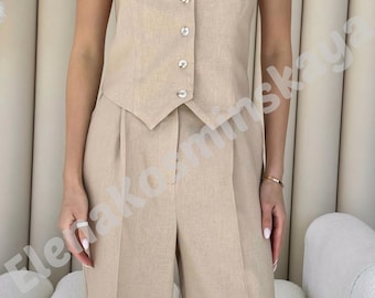 May include: A beige sleeveless vest with a V-neck and matching wide-leg pants. The vest has a button-up closure with five buttons. The outfit is made of a light-colored fabric, suitable for a business or casual setting.