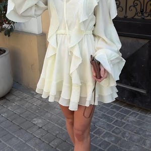 May include: A pale yellow mini dress featuring long, voluminous bell sleeves, a tiered ruffled skirt, and a high neckline with a tie closure. The dress has a cinched waist and is paired with a small brown leather crossbody bag and olive green shoes.