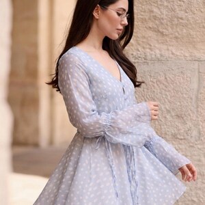 May include: A pale blue dress with white polka dots. The dress has long, sheer sleeves with frilled cuffs and a V-neck. A tie defines the waist, and the skirt flares. The dress is made of a lightweight material.