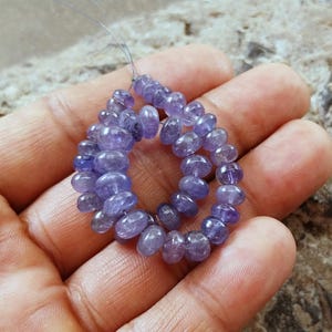 Könnte beinhalten: Eine Strähne aus violetten und blauen Edelsteinperlen, wahrscheinlich Amethyst oder Tansanit, auf einem dünnen Draht aufgefädelt. Die Perlen sind rund und glatt, mit einer leichten Größenvariation. Die Perlen sind in einem lockeren Kreis angeordnet, wobei die Enden des Drahtes sichtbar sind.