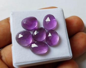 May include: Six faceted, oval amethyst gemstones in a white box. The gemstones are a deep purple colour with a polished, reflective surface. Each stone has a unique cut, catching the light and creating a sparkling effect. The gemstones are displayed on a white background.