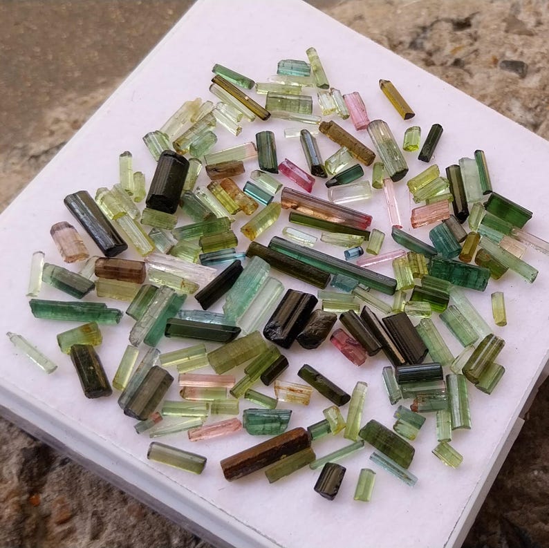 Pu&ograve; includere: Una collezione di cristalli di tormalina verde, nera e rosa di varie dimensioni e forme. I cristalli sono tutti tagliati in lunghi prismi rettangolari sottili.
