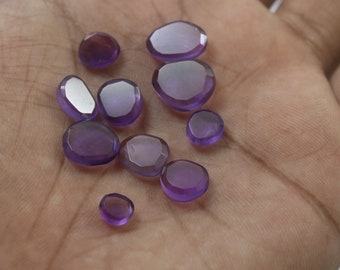 May include: A collection of ten purple amethyst gemstones, each with a unique, faceted shape. The stones are arranged on a hand, showcasing their various sizes and cuts.