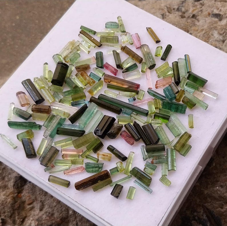 Pu&ograve; includere: Una collezione di punte di gemme di turmalina verde, nera e rosa. Le punte sono di varie dimensioni e forme e sono disposte su una superficie bianca.
