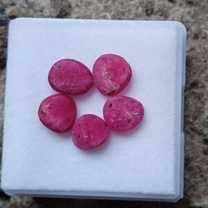 May include: Six pink gemstones in a variety of shapes and sizes are displayed on a white surface. The gemstones are smooth and have a slight sparkle.