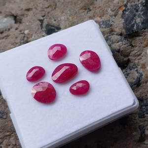 May include: Six faceted, oval-shaped, deep red gemstones are displayed on a white surface. The gems vary in size, with a brilliant cut that reflects light. The stones are likely rubies, showcasing a rich, saturated color.