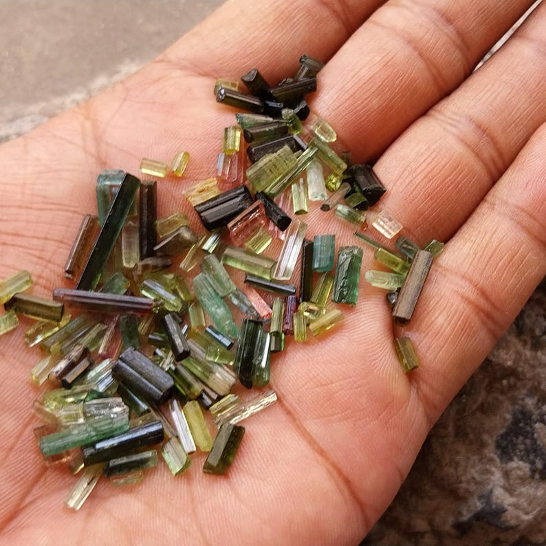 Pu&ograve; includere: Una collezione di piccoli cristalli di tormalina verde, nero e marrone di varie forme e dimensioni. I cristalli sono disposti in un mucchio sulla mano di una persona.