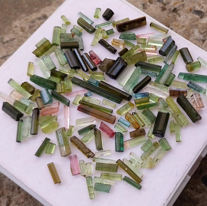 Pu&ograve; includere: Una collezione di perline di gemme di turmalina verde, nera e rosa. Le perline sono tutte tagliate a forma di cilindri lunghi e sottili e sono disposte in uno schema casuale su una superficie bianca.
