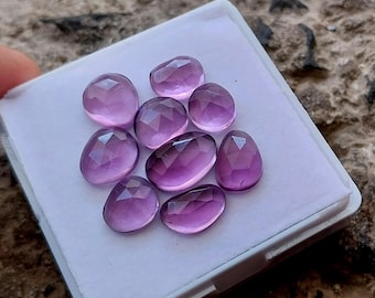 May include: Nine faceted amethyst gemstones displayed in a white tray. The gemstones are various shapes, including oval and teardrop cuts. The colour is a deep purple, with lighter shades in the facets.