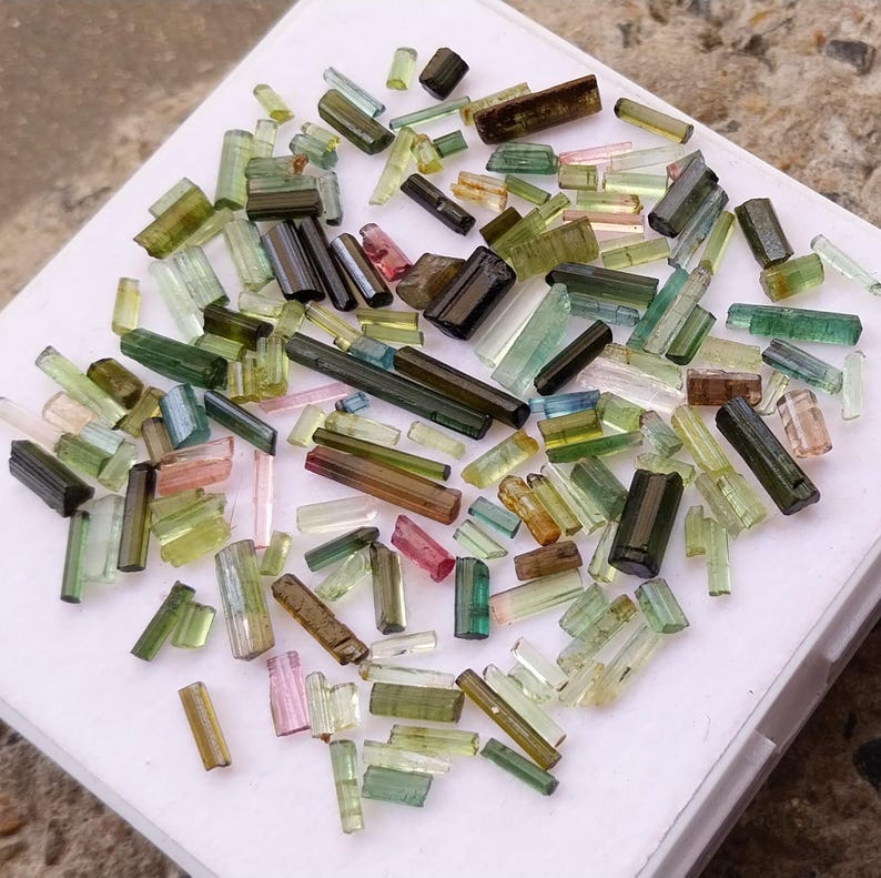 Pu&ograve; includere: Una collezione di piccoli cristalli di tormalina traslucidi e colorati in varie tonalit&agrave; di verde, rosa e blu. I cristalli sono disposti su una superficie bianca.