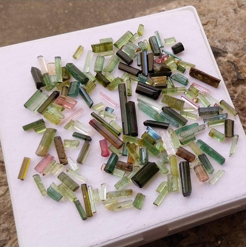 Pu&ograve; includere: Una collezione di piccoli cristalli di tormalina traslucidi e colorati in varie tonalit&agrave; di verde, rosa e blu. I cristalli sono disposti su una superficie bianca.