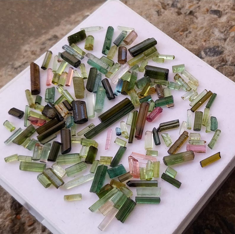 Pu&ograve; includere: Una collezione di cristalli di turmalina verde, blu e rosa di varie dimensioni e forme. I cristalli sono disposti su una superficie bianca.