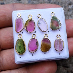 Könnte beinhalten: Acht Edelsteinanhänger mit goldfarbenen Fassungen werden auf einer weißen Oberfläche präsentiert. Die Edelsteine variieren in der Farbe, darunter Rosa, Grün und Klar. Jeder Anhänger hat oben eine Schlaufe zum Befestigen an einer Kette oder einem anderen Schmuckbestandteil.