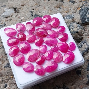 May include: A collection of faceted, oval-shaped, and round ruby gemstones in varying sizes. The gemstones are a vibrant pinkish-red color and are arranged on a white tray. The tray is set against a textured, gray background.
