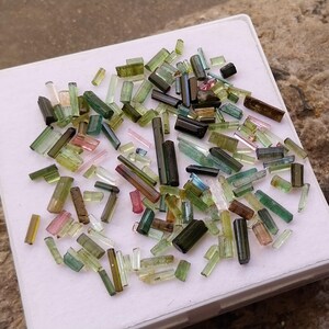 Pu&ograve; includere: Una collezione di punte di turmalina gemma verde, nera e rosa. Le punte sono di varie dimensioni e forme e sono disposte su una superficie bianca.