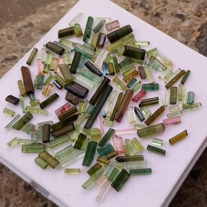 Pu&ograve; includere: Una collezione di cristalli di tormalina verde, blu e rosa di varie dimensioni e forme. I cristalli sono disposti su una superficie bianca.