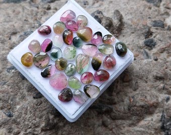 May include: A collection of small, faceted, multi-coloured tourmaline gemstones arranged on a white square display pad. The stones feature various shades of pink, green, yellow, and clear tones, each with a polished, reflective surface and unique shape.