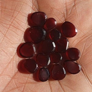 May include: A collection of small, deep red, faceted gemstones with a polished finish. These loose stones feature a rounded, irregular shape and are arranged in a cluster, displaying a translucent quality that catches the light across their surfaces.