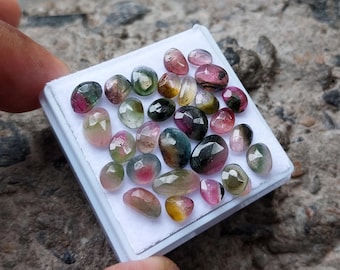 May include: A collection of small, faceted, multi-coloured tourmaline gemstones arranged on a white display pad. The stones feature various shades of pink, green, yellow, and black, with polished surfaces that reflect light in a variety of patterns.