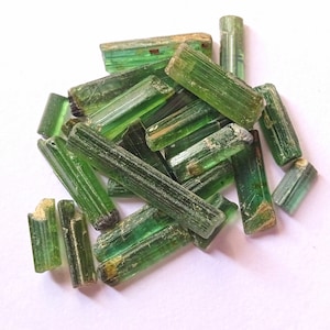 Op de afbeelding: Een verzameling groene, ruwe, ongeslepen, ruwe, natuurlijke edelsteenkristallen. De kristallen zijn lang en slank, met een gestreepte textuur. Ze zijn op een witte achtergrond in een stapel gerangschikt.