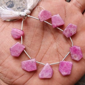 May include: A strand of eight irregular, faceted pink ruby gemstones strung on a thin silver-colored wire. The strand is finished with a white tassel at the top, featuring a metallic silver-toned cap that secures the wire and the tassel together.