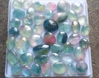 May include: A collection of over 40 loose, natural gemstone cabochons in a variety of colours, including pink, green, blue, and clear. The stones are all oval shaped and have a smooth, polished finish.