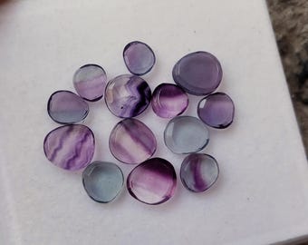 May include: A collection of polished fluorite gemstones in various shapes and sizes. The stones display a range of colors, including translucent lavender, purple, and hints of blue. The gemstones are arranged on a white surface.