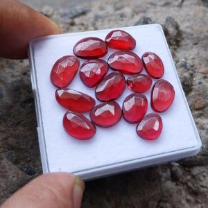 Può includere: Una collezione di tredici pietre preziose sfaccettate, di colore rosso intenso e forma ovale, disposte su un vassoio espositore quadrato bianco. Ogni pietra presenta una superficie lucida con molteplici sfaccettature geometriche.