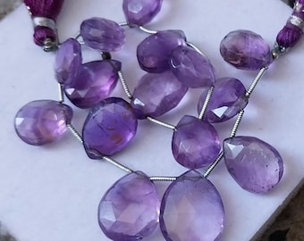 May include: A strand of purple amethyst gemstone beads, each bead is faceted and shaped like a teardrop. The beads are strung on a silver wire.