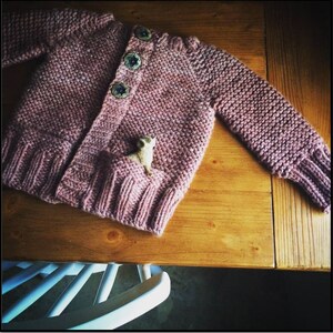 May include: A pink knitted cardigan with a small wooden animal figurine in the pocket. The cardigan has a button closure and a textured knit pattern.