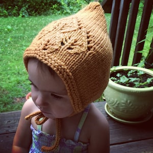 May include: A child wearing a knitted yellow bonnet with a lacy pattern. The bonnet has a long tie that is tied under the chin.