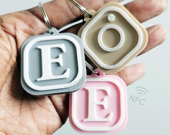May include: Three keychains with a silver ring. The first keychain is gray with a white letter E. The second keychain is pink with a white letter E. The third keychain is beige with a white circle inside a square.