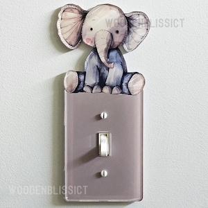 May include: A light switch cover with a cute cartoon elephant illustration. The elephant is sitting on top of the cover and is wearing a blue shirt. The cover is a light gray color.