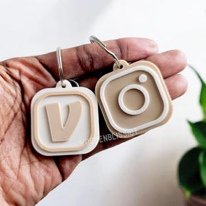 May include: Two keychains, one with a white letter V and the other with a brown Instagram logo, both with white borders.