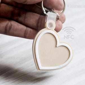 Pu&ograve; includere: Un portachiavi a forma di cuore beige con un contorno bianco e un anello argentato. Il portachiavi ha un chip NFC e il testo "NFC" &egrave; stampato sul retro.