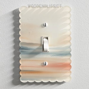 Op de afbeelding: Een lichtschakelaar-afdekplaat met een gegolfde rand en een aquarel-ontwerp in blauwe, bruine en perzikkleurige strepen. De schakelaar staat in de uit-stand. De tekst "WOODENBLISSICT" is bovenaan de afdekplaat gedrukt.