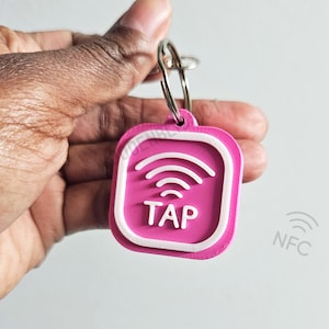 May include: A pink keychain with a white border and a white wireless symbol with the word "TAP" in white letters. The keychain has a silver ring attached to it.