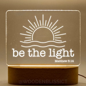 Könnte beinhalten: Ein weißes, leuchtendes Acrylschild mit einem Sonnen- und Wellenmotiv und dem Text "be the light" mit dem Bibelvers "Matthäus 5:14". Das Schild steht auf einem Holzsockel.