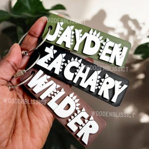May include: Three personalized keychains with the names Jayden, Zachary, and Wilder. Each keychain is made of a different color of leather and features a white dinosaur design.
