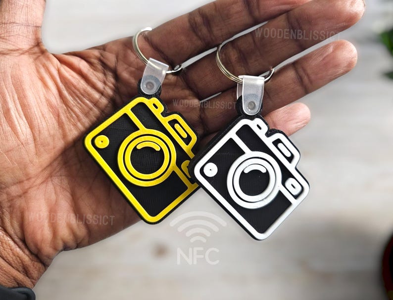 Pu&ograve; includere: Due portachiavi a forma di macchina fotografica con disegni in bianco e nero. Il portachiavi a sinistra ha un contorno giallo e il portachiavi a destra ha un contorno bianco. Entrambi i portachiavi hanno la scritta "NFC" stampata su di essi.
