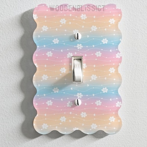 Puede incluir: Una tapa de interruptor de luz con un fondo degradado de arcoíris pastel y un patrón floral blanco. La tapa del interruptor tiene un borde festoneado.