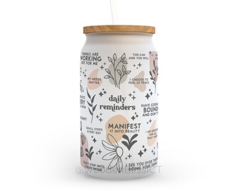 May include: White reusable tumbler with a wooden lid and a straw. The tumbler has a black and white design with pink accents and inspirational quotes like "daily reminders" and "manifest it into reality".