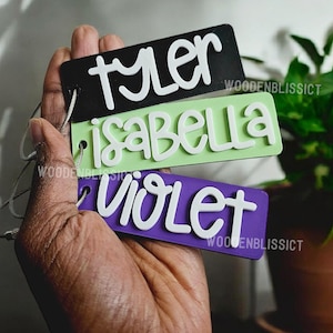 May include: Three wooden name tags with black, green, and purple backgrounds. The names "Tyler", "Isabella", and "Violet" are written in white on the tags.