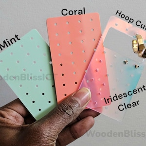 May include: Three earring display cards in mint green, coral, and iridescent clear. The iridescent card has a cutout for hoop earrings. The text "Hoop Cutout" is printed on the card. The text "Iridescent Clear" is printed on the card. The text "Coral" is printed on the card. The text "Mint" is printed on the card.